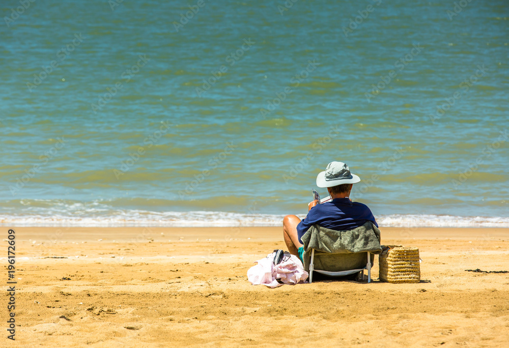 Senior man reading a newspaper on the beach at the water's edge. Senior man reading a newspaper on the beach at the water's edge.