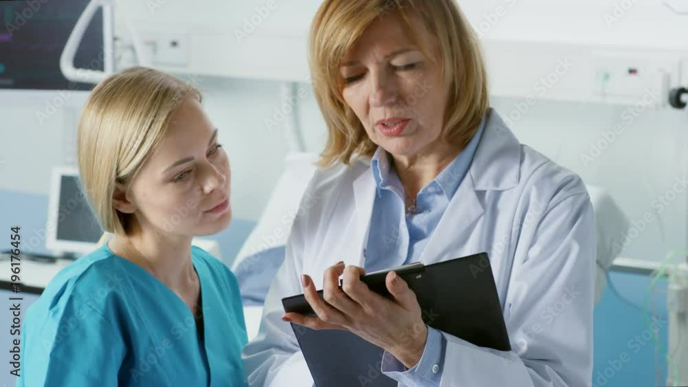 Professional Female Doctor and Young Nurse Discuss Patient's Chart. In the Background Sick Man Lying in the Bed Waiting to be Cured. Shot on RED EPIC-W 8K Helium Cinema Camera. Professional Female Doctor and Young Nurse Discuss Patient's Chart. In the Background Sick Man Lying in the Bed Waiting to be Cured. Shot on RED EPIC-W 8K Helium Cinema Camera.