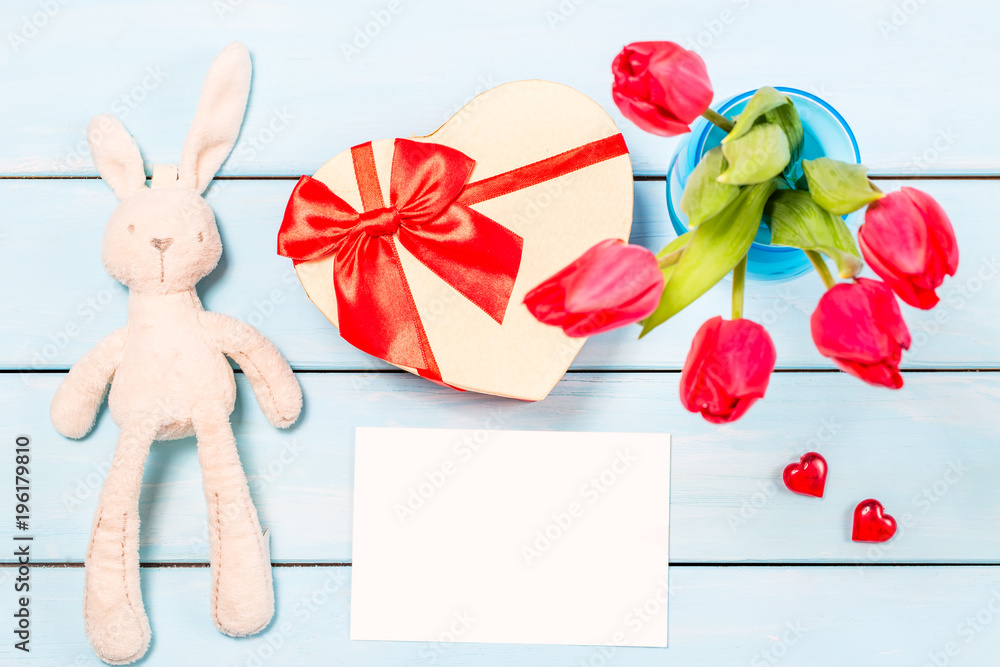 Colorful red spring tulip flowers in nice blue vase, blank photo frame and stuffed toy bunny with decorative heart on light wooden background as greeting card. Mothersday or spring concept. Top view