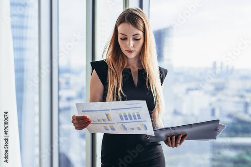 Female financial analyst holding papers studying documents standing against window with city view