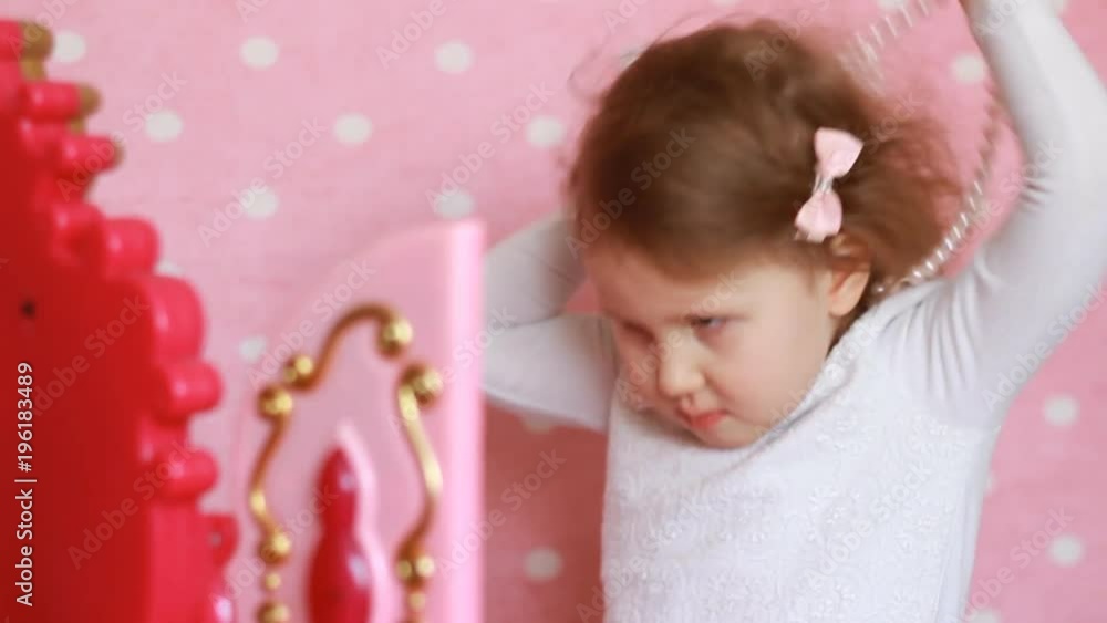 Beautiful little girl dresses up, puts on beads and looking in the mirror. A cute child fashionista plays a real lady. Pink glamor background and children's chest of drawers