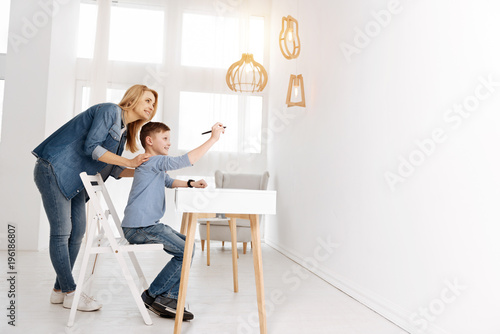 Futuristic development. Nice delighted pleasant woman standing behind her son and holding his shoulders while looking at him using new technology