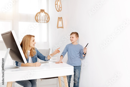 Intelligent boy. Delighted happy young woman smiling and listening to her son while sitting at the table