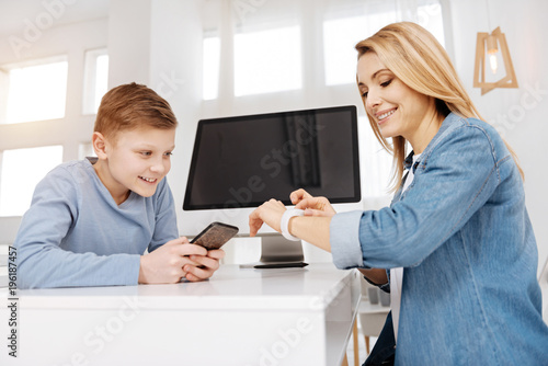 Modern generation. Happy nice young woman smiling and holding his smartphone while looking at his mothers smartwatch