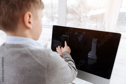 Fototapeta Naklejka Na Ścianę i Meble -  Digital device. Smart cute positive boy sitting in front of the digital screen and holding a smartphone while using this device