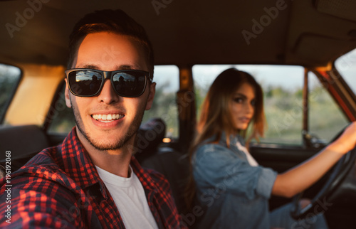Horizontal portrait of happy romantic young couple going on a long drive in car on sunset light. Handsome young man wearing sunglasses with his beautiful girlfriend on a road trip. Travel concept.