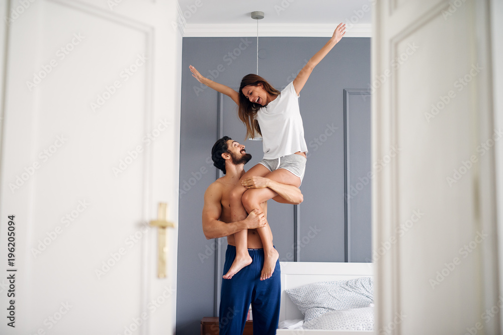 Strong shirtless hipster man holding his adorable wife with raised arms on a shoulder in the bedroom.