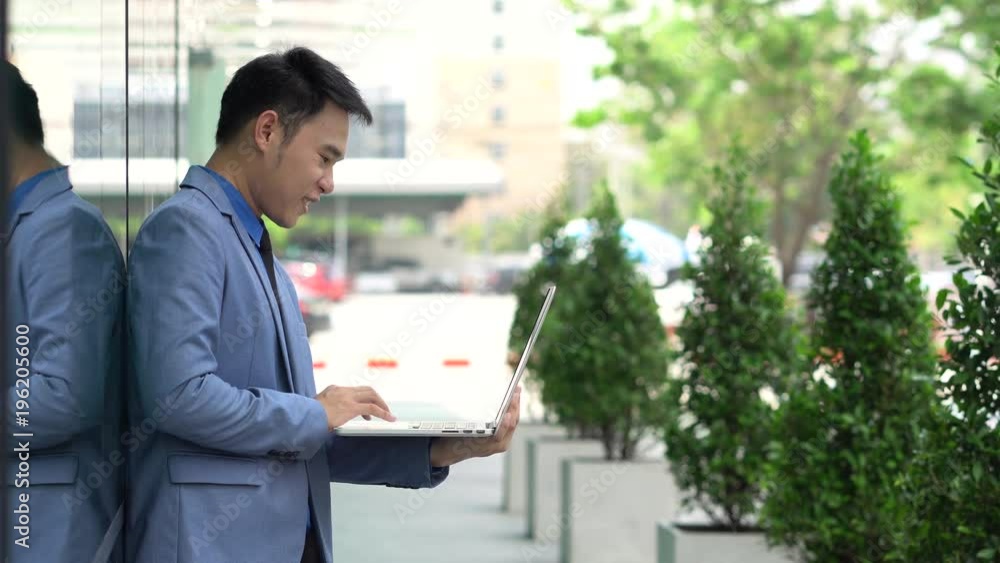 Asian businessman casual using laptop standing on floor beside modern office with happiness on a summers day Asian businessman casual using laptop standing on floor beside modern office with happiness on a summers day
