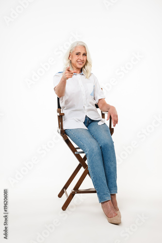 Full length portrait of joyful mature woman 70s with gray hair sitting on wooden chair and pointing finger on camera, isolated over white background