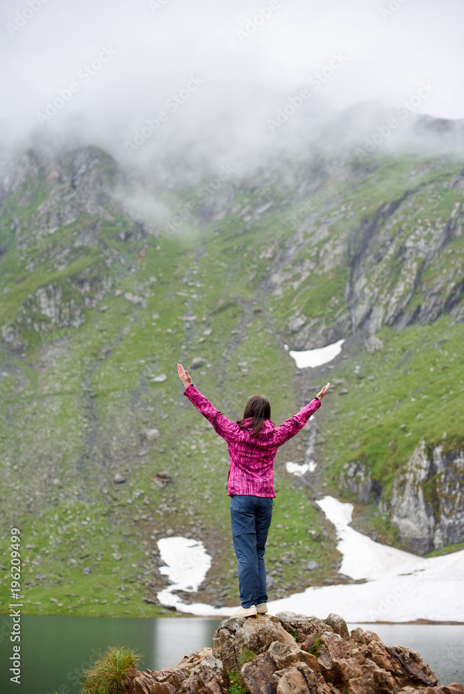 Woman is standing with her back on a rock with her arms wide open and looks up against background of mighty mountains and the lake at the foot, Transylvania, Romania. Woman is standing with her back on a rock with her arms wide open and looks up against background of mighty mountains and the lake at the foot, Transylvania, Romania.