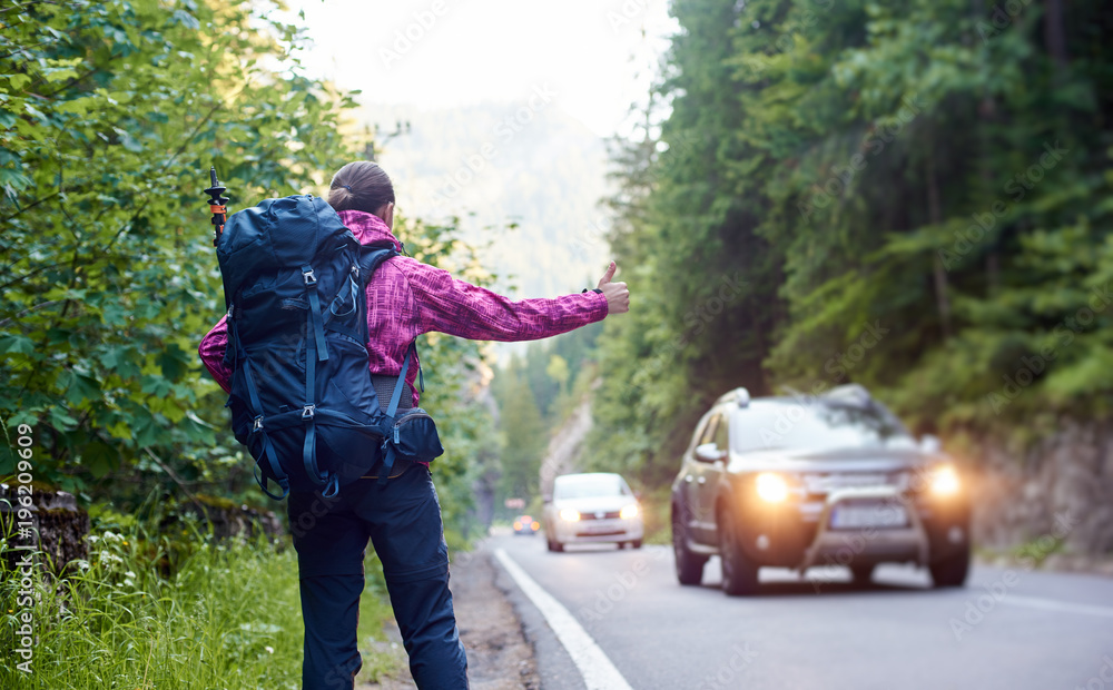 Φωτογραφία Female tourist with backpack hitchhiking a car on mountain road with green rocky hills and trees near