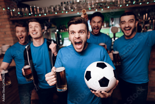 Five soccer fans drinking beer celebrating and cheering at sports bar.