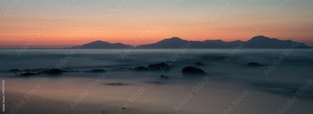 A long exposure of the sea at golden hour, as the sun sets behind the mountains. The sea looks ethereal as it breaks over the rocks and beach.