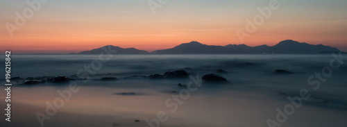 A long exposure of the sea at golden hour  as the sun sets behind the mountains. The sea looks ethereal as it breaks over the rocks and beach.