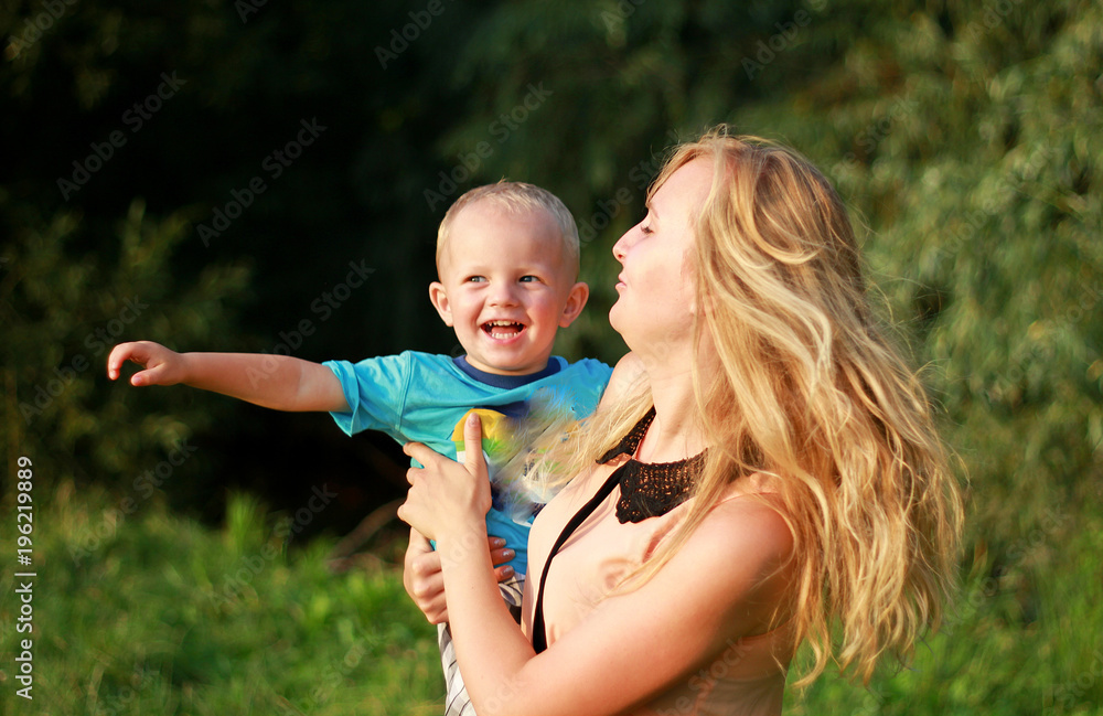 Fototapeta premium Young blonde mother in playing with her child in the green lawn. Happy fun summer photo. Smiling laughing boy with his mom. Sunny day outdoors in nature. Warm hug and embrace.