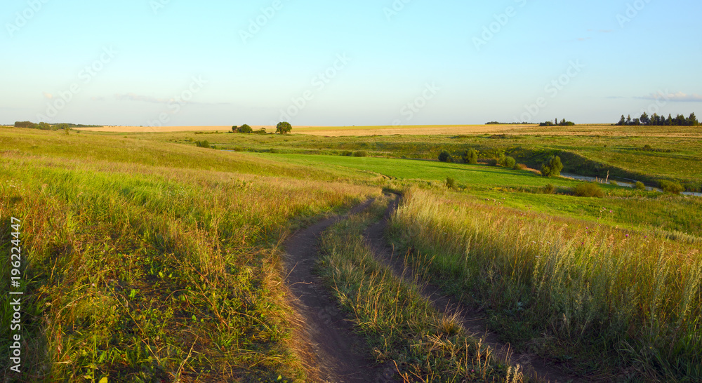 Obraz premium Sunny summer landscape with ground country road.Beautiful view.Green hills,fields and meadows.Tula region,Russia.Sunset.Quiet weather.Calm.Warm sunlight.Cloudless clear blue sky.