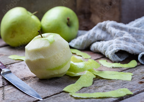 Green peeled apple on wooden desk with knife and apples behind.