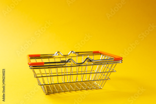 Close up of metal grocery basket for shopping in supermarket with lowered handles and orange plastic elements isolated on yellow background. Concept of shopping. Copy space for advertisement photo