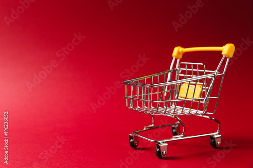 Close up of supermarket grocery push cart for shopping with black wheels and yellow plastic elements on handle isolated on red background. Concept of shopping. Copy space for advertisement