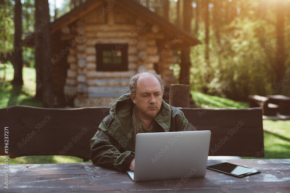 An adult serious partly bald man entrepreneur is working on the laptop during his vacations while sitting outdoors in a forest, with small shack behind and the forest in a defocused background An adult serious partly bald man entrepreneur is working on the laptop during his vacations while sitting outdoors in a forest, with small shack behind and the forest in a defocused background