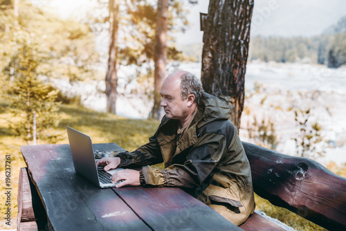An adult partly bald game warden in the overalls of marsh color is sitting outdoor on the bench at the big wooden table and having a conversation with his family via video call from laptop