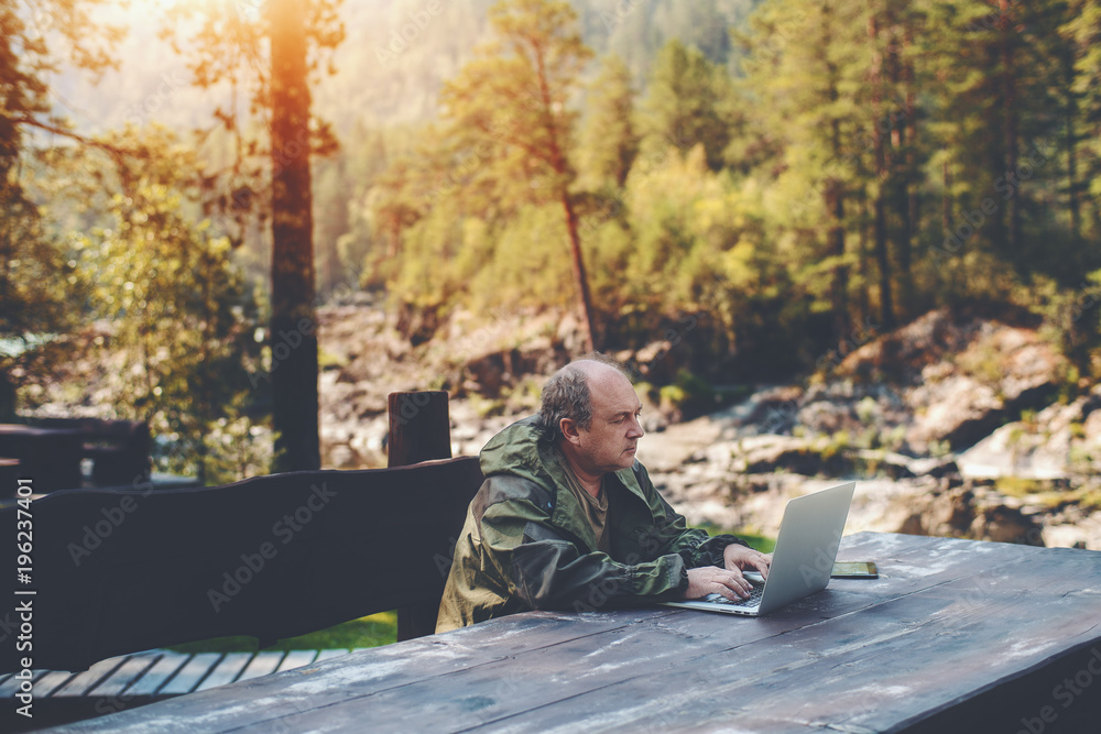 Elderly man employer is spending his vacations with work outdoors: sitting with the laptop at the wooden table with a digital tablet next to him and summer pine forest in a defocused background Elderly man employer is spending his vacations with work outdoors: sitting with the laptop at the wooden table with a digital tablet next to him and summer pine forest in a defocused background