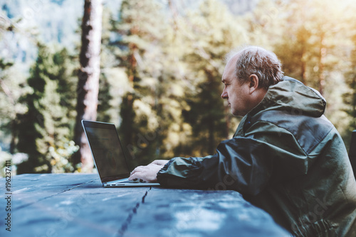 Side view of an aged male forester in dark-green coverall making a report on his laptop while sitting outdoor at the wooden table with the forest in a blurry background photo