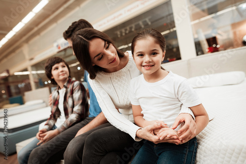 Young beautiful woman sits on orthopedic mattress with cheerful boy and girl in furniture store.