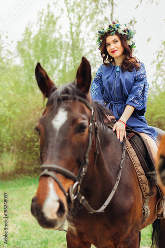 cute girl in a beautiful blue dress sitting on a horse. big flower wreath on head of nice woman