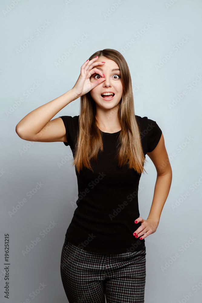 Positive emotions and feelings on white background.Caucasian female with long hair, wearing black T-shirt, looking at the camera through fingers in okay gesture. Face expressions Positive emotions and feelings on white background.Caucasian female with long hair, wearing black T-shirt, looking at the camera through fingers in okay gesture. Face expressions