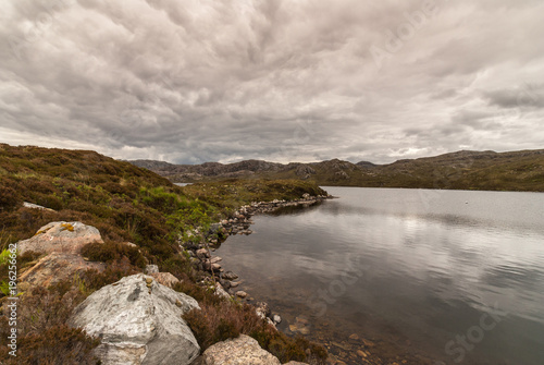 Loch Tollaidh, Scotland - June 9, 2012: Heavy dark brown cloudscape over lake colors water dark. Set between rocky mountains. Green grass and rocks in front. Wide landscape shot.