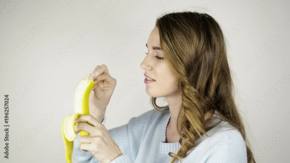 Seductive fair haired woman in gray peeling a banana and biting it. She is looking at you. Locked down real time establishing shot Seductive fair haired woman in gray peeling a banana and biting it. She is looking at you. Locked down real time establishing shot