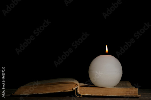 White burning candle with opened book on black background