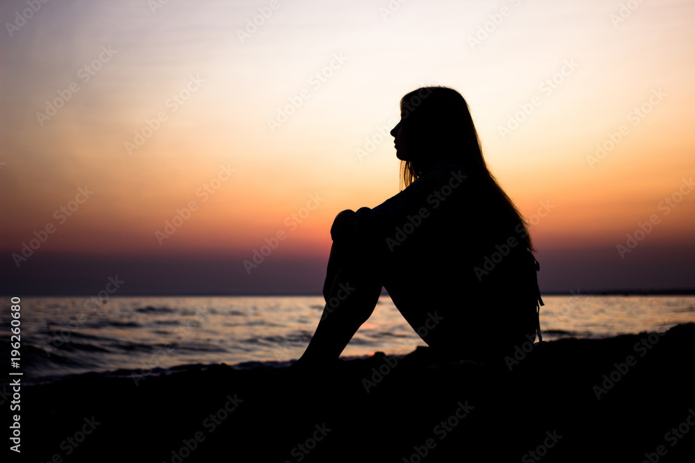 Silhouette of a young girl sitting on the sand overlooking the beach with the sun as a background. sunset