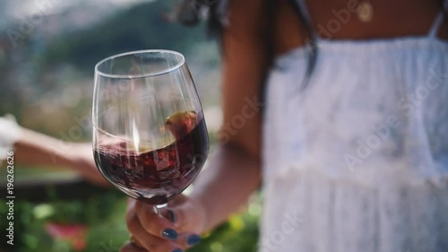 Hand of young unrecognizable woman in white dress shakes glass of red wine outdoors on hot summer sunny day photo