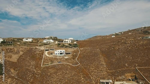 Black car moving up the road to the hill with luxury white villas on top. Greek Cyclades with dry land, brown stones and arid bushes. Faros Armenistis, Mykonos island, Greece, Aerial.