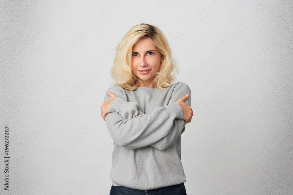 Portrait of young blonde girl embrace herself, wears casual gray sweater and feels comfortable and warm. Positive emotions. Isolated over white wall Portrait of young blonde girl embrace herself, wears casual gray sweater and feels comfortable and warm. Positive emotions. Isolated over white wall