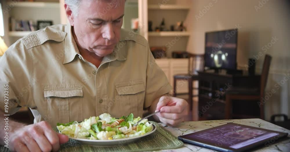 A man sitting at home eating a salad of leafy greens for lunch while he views something on his IPAD. A man sitting at home eating a salad of leafy greens for lunch while he views something on his IPAD.