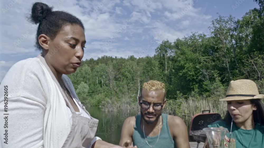 Group of young diverse people having lunch together at table by lake, women helping others with serving Group of young diverse people having lunch together at table by lake, women helping others with serving