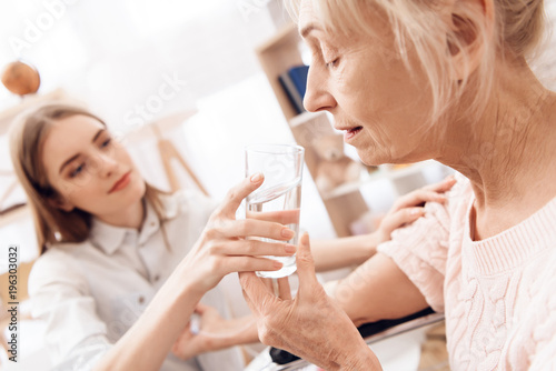 Girl is nursing elderly woman at home. Girl is helping woman with glass of water.