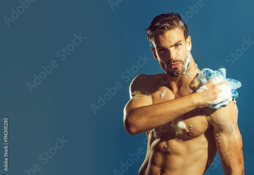 Bodybuilder, sexy strong man washing with foam sponge after workout. Man with bristle and concentrated face take shower in bathroom. Advertising for men's cosmetics for body care. Copy space.