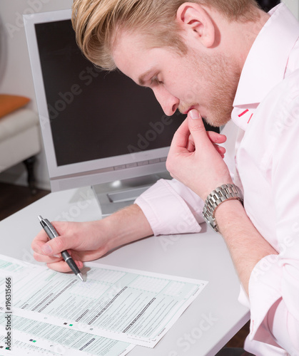 Junger Mann sitzt am Schreibtisch und arbeitet an Steuerformularen photo