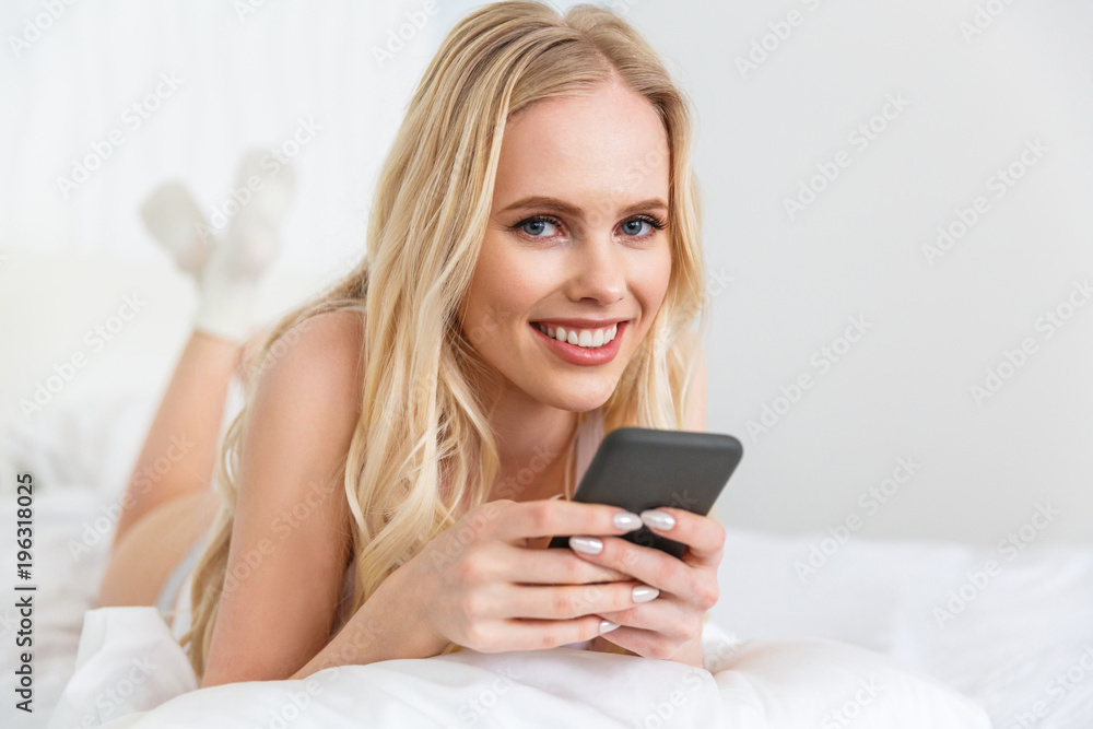 beautiful young blonde woman holding smartphone and smiling at camera while lying on bed beautiful young blonde woman holding smartphone and smiling at camera while lying on bed