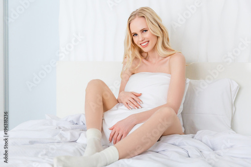 beautiful young blonde woman sitting on bed and smiling at camera