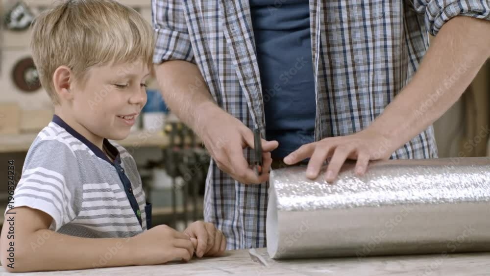 Man making crafts with son, cutting piece of Styrofoam roll covered with foil, child taking it and smiling Man making crafts with son, cutting piece of Styrofoam roll covered with foil, child taking it and smiling