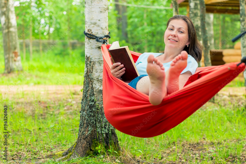 Naklejka premium happy 30 year old woman reading a book, sitting in a red hammock in the garden