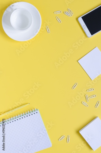 Top view office desk with notepads, phone, paper for notes, coffee cup on yellow background.