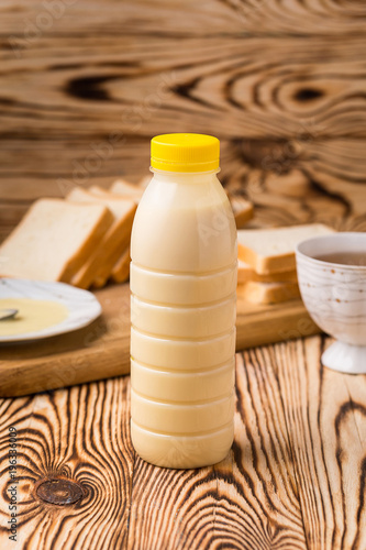 A bowl and a bottle of condensed milk and a teaspoon on a wooden table with background of toasts and tea cup