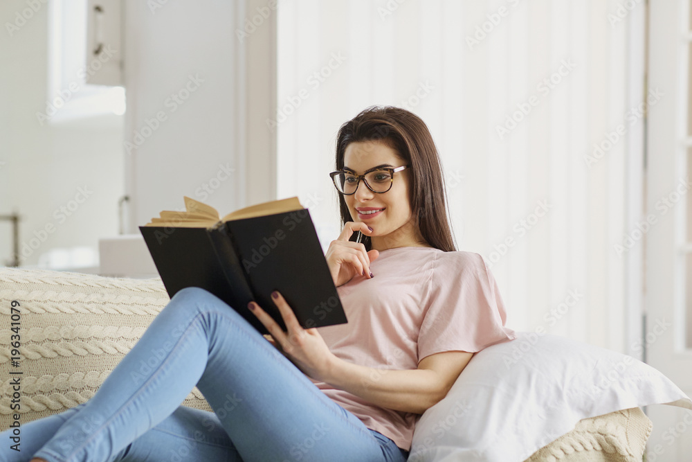 A girl in glasses reads an interesting book sitting on the couch in comfort in the room. A girl in glasses reads an interesting book sitting on the couch in comfort in the room.