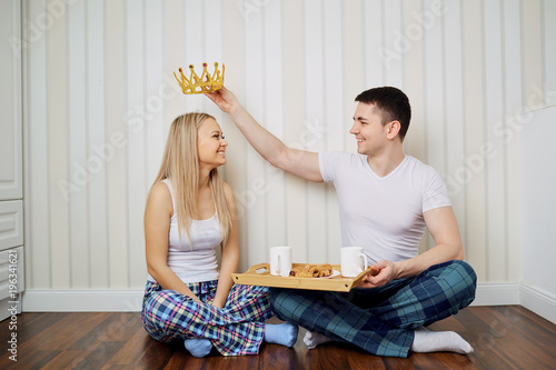 A happy couple in pajamas sit on the floor in a room against a striped wall background.